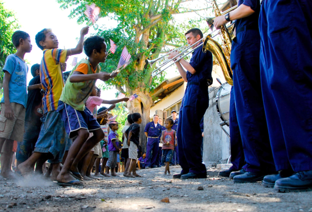 Gruppe von Kindern, die Musikinstrumente im Freien spielen, mit einer Menge, die Fahnen schwenkt, im Hintergrund, Bäume, Gebäude und einen klaren blauen Himmel.