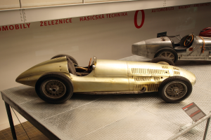 Zwei Oldtimer-Rennwagen in einem Museum ausgestellt, mit einem Textschild im Vordergrund und einer Textwand im Hintergrund.