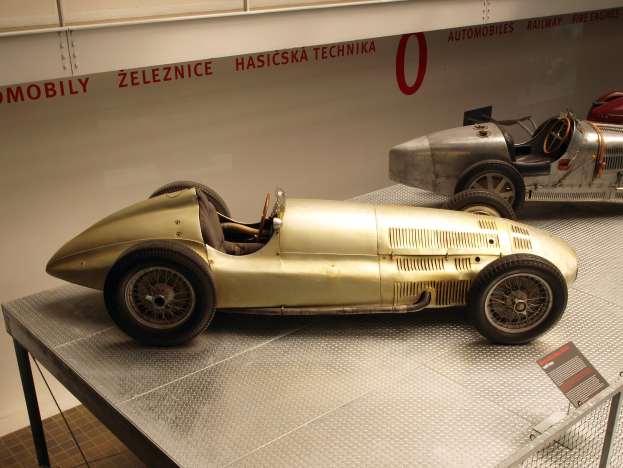 Zwei Oldtimer-Rennwagen in einem Museum ausgestellt, mit einem Textschild im Vordergrund und einer Textwand im Hintergrund.