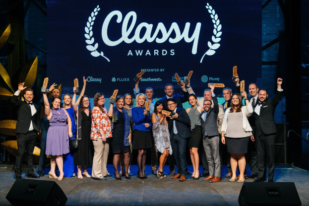 Gruppe von Menschen auf einer Bühne, die Preise halten, mit Lautsprechern darunter und einem Bildschirm im Hintergrund, der "Classy Awards 2019" anzeigt.