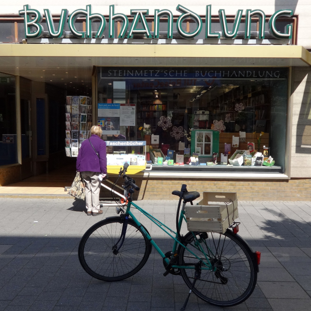 Eine Frau steht neben einem Fahrrad, das vor einem Geschäft geparkt ist, mit einem Fahrradrad in Vordergrund und Bücher durch die Glasscheibe des Geschäfts sichtbar.