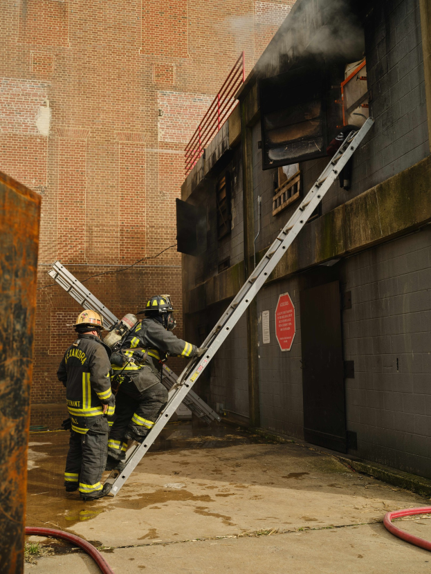 Feuerwehrleute mit Helmen klettern an einer Leiter zu einem raucherfüllten Gebäude hoch, mit einem Metallobjekt links und Rohren auf dem Boden darunter.