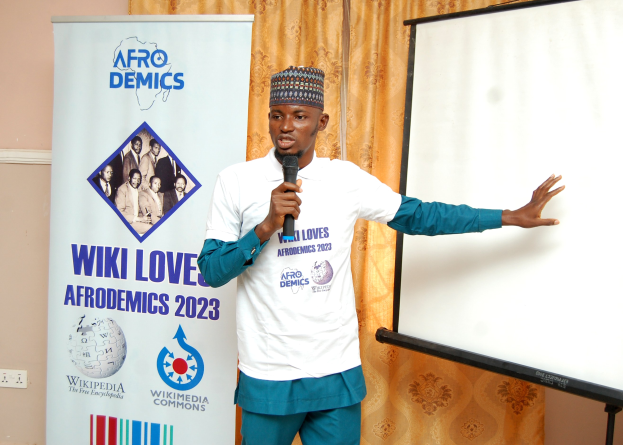 Mann in einer Mütze, der ein Mikrofon hält und vor einer Whiteboard, einer Schalttafel, einem Banner und einem Vorhang spricht.