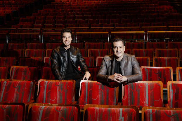 Zwei Männer in schwarzen Jacken, die während einer 'Dancing with the Stars'-Show in einer roten Auditoriumsreihe sitzen und lächeln.