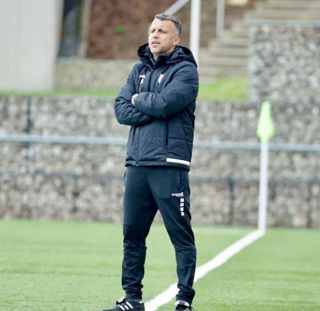 Ein Fußballtrainer auf einem Feld mit verschränkten Armen, der eine schwarze Jacke und Hose trägt, vor einem Zaun, Treppe und Wand.