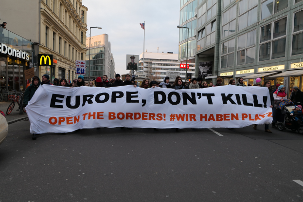 Eine Gruppe von Menschen marschiert mit einem Banner, auf dem 'Europa, töte nicht, öffne die Grenzen' steht, die Straße entlang, mit einem geparkten Auto auf der linken Seite, Gebäuden und einer Flagge im Hintergrund unter einem klaren blauen Himmel.