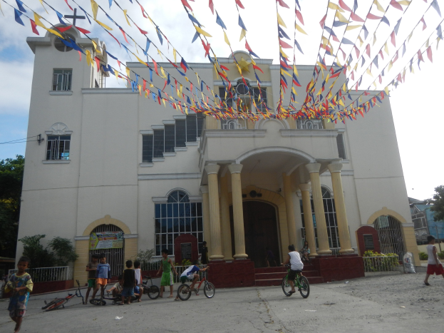 Eine Gruppe von Menschen auf Fahrrädern vor einer weißen Kirche mit architektonischen Details wie Fenstern, Säulen, Bögen, Türen und einem Kreuzsymbol, vor einem Hintergrund aus Pflanzen, einem Zaun, einer Fahne, Bäumen und einem bewölkten Himmel, mit bunten Fahnen, die an einem Seil befestigt sind.