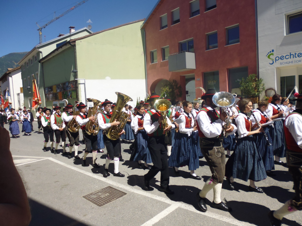 Eine Gruppe von Menschen in traditioneller bayrischer Tracht, die auf der Straße Musikinstrumente spielen und durch eine Straße mit Gebäuden gehen, einige halten Fahnen, mit einem Hügel und einem klaren blauen Himmel im Hintergrund.