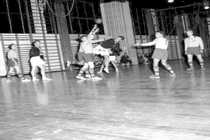Eine Gruppe von Männern, die Handball auf einem Holzboden mit einer Wand und Lampen im Hintergrund spielen, in Schwarz-Weiß dargestellt.