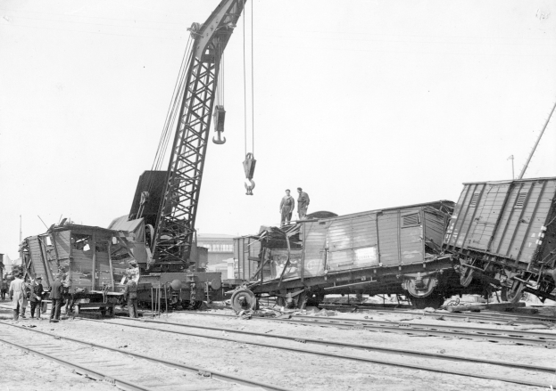 Schwarzes und weißes Bild einer Zugkatastrophe mit einem Kran, der ein Waggon auf die Schienen hebt, Menschen in der Nähe, Gebäude und einen klaren Himmel im Hintergrund.