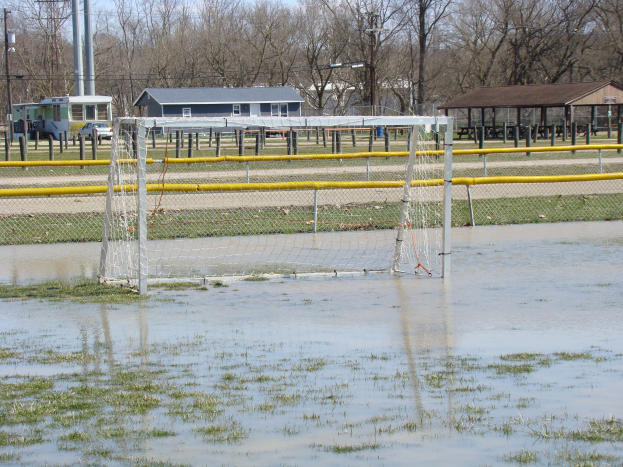 Fußballtor in einem überfluteten Feld mit umliegender Grasfläche und Wasser, Schuppen, Pfosten, Bäumen, Fahrzeugen und einem klaren blauen Himmel im Hintergrund.