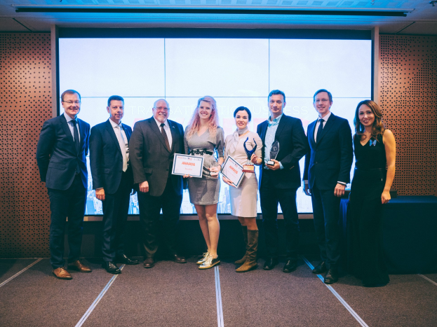 Gruppe von strahlenden Menschen, die Zertifikate halten und vor einem Bildschirm stehen, mit einem Tisch und einem wandmontierten Objekt im Hintergrund, Gewinner der International Business Awards 2018.