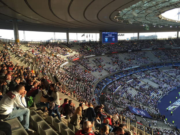 Große Menschenmenge in einem Stadion bei einem Fücballspiel, mit einer Bühne auf der rechten Seite, Fahnen, Masten, einem Bildschirm und der Allianz Arena in München, Deutschland im Hintergrund.