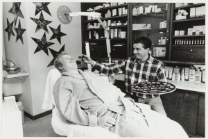 Ein Mann lächelt im Zahnarztstuhl, während ein anderer Mann neben ihm steht und ein Tablett hält, mit einem Waschbecken, Sternen, einer Uhr und Regalen mit Flaschen im Hintergrund.