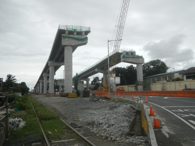 Baustelle mit einer Brücke im Hintergrund, Straße durch Verkehrskegel markiert, Eisenbahnschiene links, verstreute Steine und Gras, Bäume und Gebäude auf beiden Seiten und ein bewölkter Himmel.