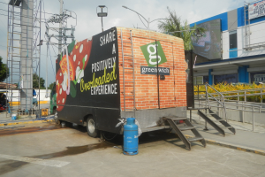 Ein Lastwagen vor einem Gebäude mit einer Tafel, auf der "Teilen Sie eine positiv uberladene Erfahrung" steht, mit Stufen, einem Zylinder am Boden, Geländern, elektrischen Masten mit Drähten, Laternenmasten, Bäumen und einem bewölktem Himmel.