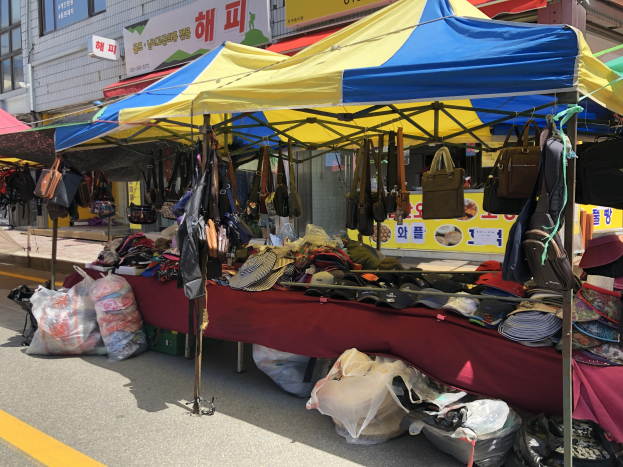 Ein Straßenmarkt mit Zelten, auf denen verschiedene Gegenstände wie Taschen und Hüte präsentiert werden, vor einem Hintergrund aus Gebäuden mit Fenstern und Schildern.