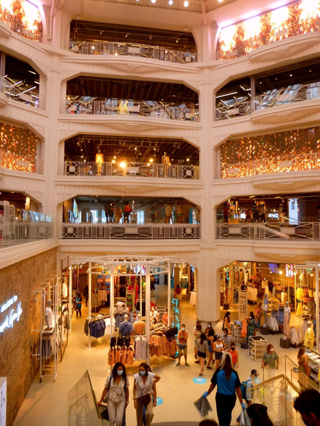 Großer Atrium eines belebten Einkaufszentrums mit Menschen, die Taschen tragen und Kleiderauslagen auf Schaufensterpuppen und Bügeln betrachten, unter Deckenlampen und Wandtexttafeln.