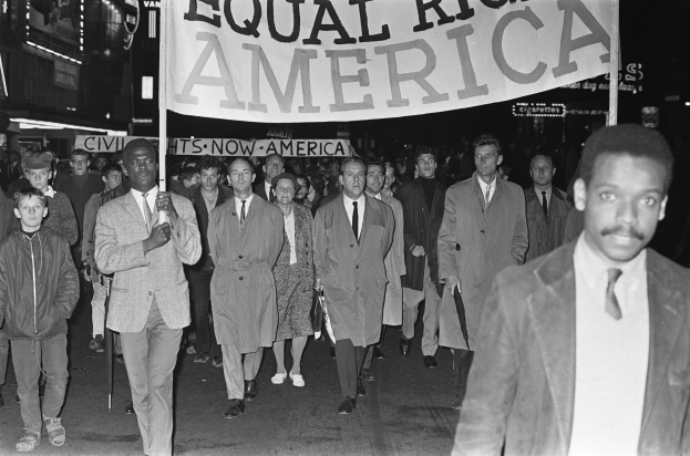 Schwarzes und weißes Foto einer Gruppe von Menschen, die mit einem Banner "Gleiche Rechte Amerika" die Straße entlang marschieren, mit Gebäuden und Straßenlaternen am Straßenrand.