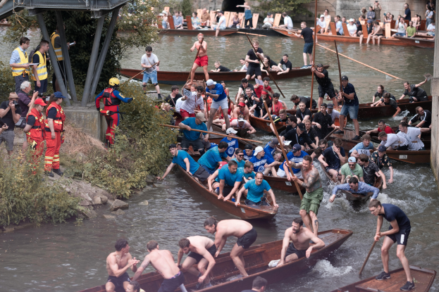 Eine Gruppe von Menschen in Kanus auf einem Fluss, einige halten Paddel, mit Pflanzen und einer Brücke links und einer Wand im Hintergrund.