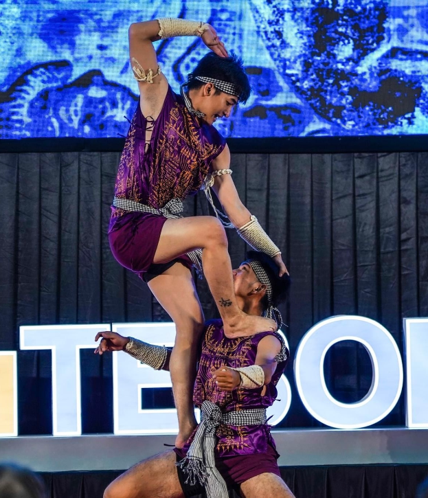 Ein Mann und eine Frau in lila Kleidern führen Akrobatik auf der Bühne durch, wobei der Mann die Frau in seinen Armen hält, während das Publikum unten zuschaut und ein schwarzer Vorhang und ein Bildschirm im Hintergrund zu sehen sind.