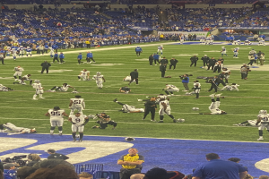 Ein Fußballspiel wird in einem Stadion mit Zuschauern gespielt, während eine Person eine Kamera hält, was auf eine Live-Übertragung hinweist.