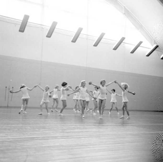 Schwarzes und weißes Bild von jungen Mädchen, die Hand in Hand tanzen und in einem Studio mit einem Ball auf der rechten Seite und Lampen an der Decke.