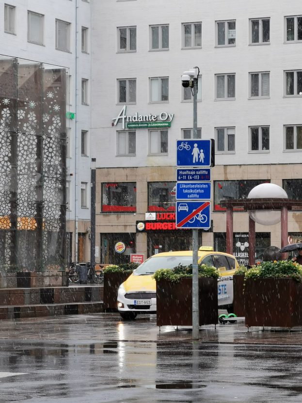 Gelbes Taxi fährt auf regennasser Stadtstraße mit einer Person mit Schirm, einem Schildständer, Blumentöpfen und einem Gebäude mit Fenstern im Hintergrund.