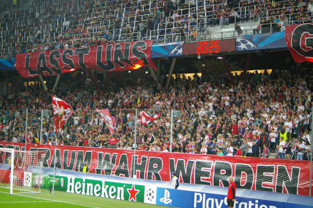 Fußballspiel in einem Stadion mit einer großen Menge an Zuschauern, saftig grünem Rasen, Torpfosten auf der linken Seite, Bodenbanner, Menschen mit Fahnen und Hintergrundgeländer, Tafeln, Lichter und andere Gegenstände.