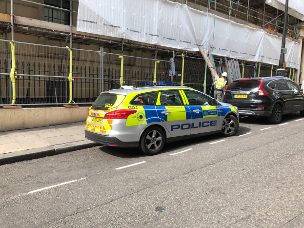 Ein Polizeiwagen steht an der Straßenseite neben einem Gebäude, mit zwei Helmträgern daneben; ein Geländer und ein Pfahl sind im Vordergrund, und ein weißes Tuch ist über das Gebäude drapiert, was auf eine Polizeiuntersuchung hindeutet.