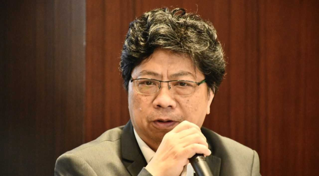 Ein Mann in Anzug und Krawatte hält ein Mikrofon und diskutiert hitzig vor einer Holzwand.