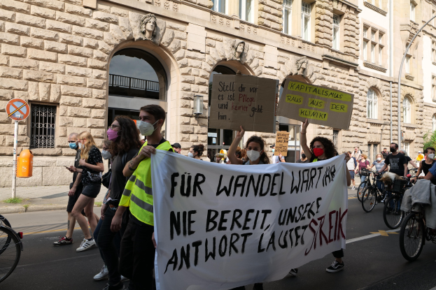 Eine Gruppe von Menschen marschiert bei einer Demonstration die Straße entlang, einige halten Schilder und andere fahren Fahrräder, im Hintergrund ist ein Gebäude mit Fenstern, Bögen, Säulen und Skulpturen zu sehen.