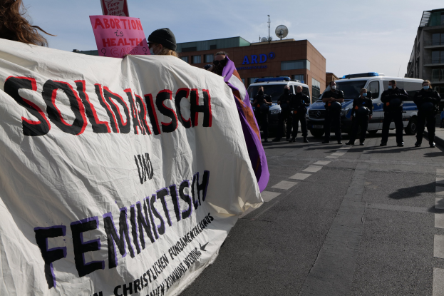Eine Gruppe von Menschen marschiert auf einer Straße, die eine Fahne mit der Aufschrift 'Solidarität und Feminismus' trägt, mit parkenden Fahrzeugen, Gebäuden, einer Schüsselantenne und einem klaren blauen Himmel im Hintergrund.