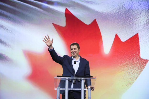 Kanadischer Premierminister Alex Salmond steht an einem Rednerpult mit Mikrofonen vor einer kanadischen Flagge, trägt ein Sakko und lächelt, mit einem sichtbaren Bildschirm im Hintergrund.