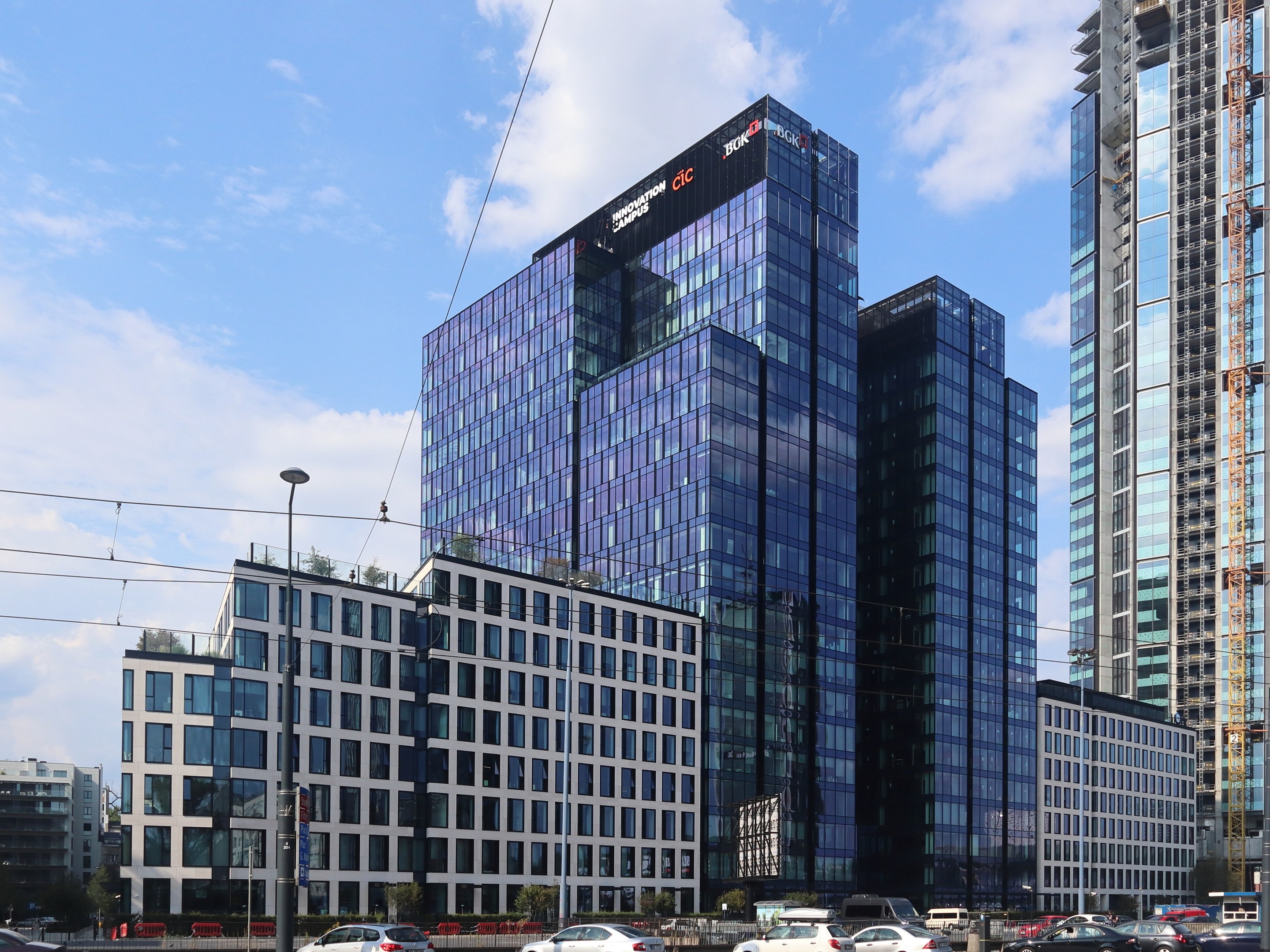 Großes modernes Gebäude mit zahlreichen Fenstern, identifiziert als Deutsche Bank Hauptsitz in Frankfurt, Deutschland, umgeben von städtischer Infrastruktur einschließlich Straßenlaternen, Fahrzeugen, Bäumen und einem bewölkten Himmel.