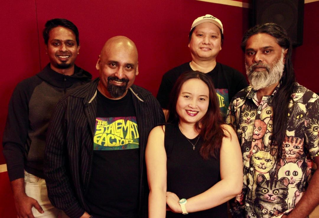 Eine Gruppe von Menschen, die zusammen in einem Aufnahmestudio stehen, lächeln und locker gekleidet sind und ihre Arme umeinander legen.