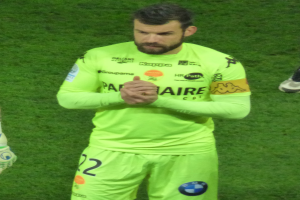 Ein Mann in einem gelben Torwarttrikot, der eine grüne T-Shirt und Shorts trägt, steht auf einem Fußballfeld und klatscht in die Hände, wobei auf der linken Seite des Bildes das Bein einer anderen Person zu sehen ist.