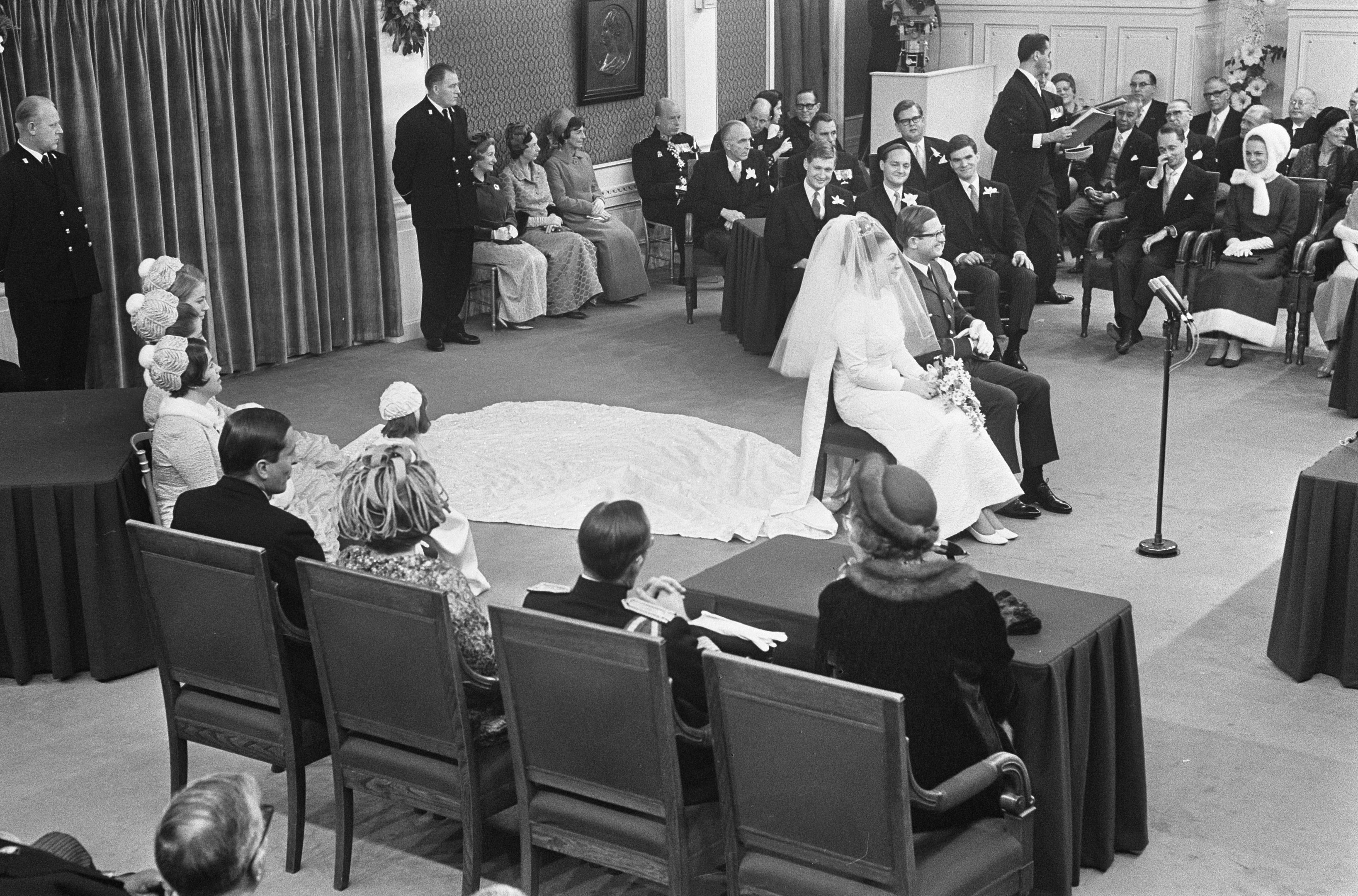 Ein Schwarz-Weiß-Foto einer Hochzeitszeremonie mit Menschen, die sitzen und stehen, einem Mann, der in ein Mikrofon spricht, einer Frau, die einen Blumenstrauß hält, einem Podium mit Mikrofon, Blumenschmuck, einem Vorhang, einem Rahmen an der Wand und einer Tür im Hintergrund.