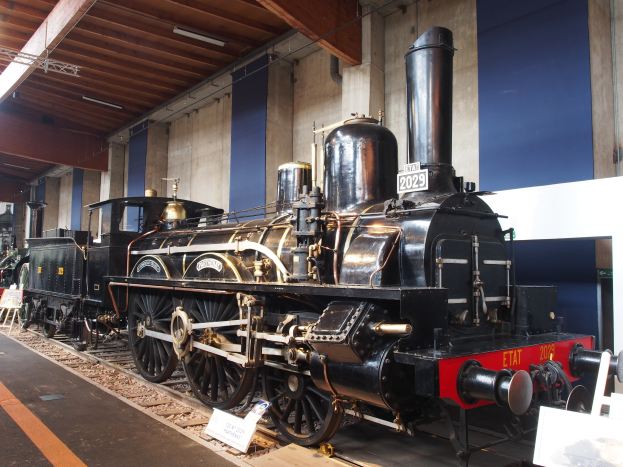 Eine große, alte Dampflokomotive in einem Museums Setting umgeben von Eisenbahnschienen, Informationsschildern und anderen Ausstellungen, mit einer Wand und beleuchteter Decke im Hintergrund.