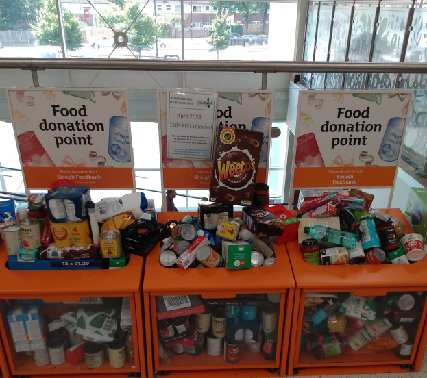 Orange Schränke mit Lebensmittelspenden wie Flaschen, Dosen und Kartons, mit Informationsschildern, durch Glasfenster betrachtet, die Fahrzeuge, Bäume, Gebäude und Himmel zeigen.