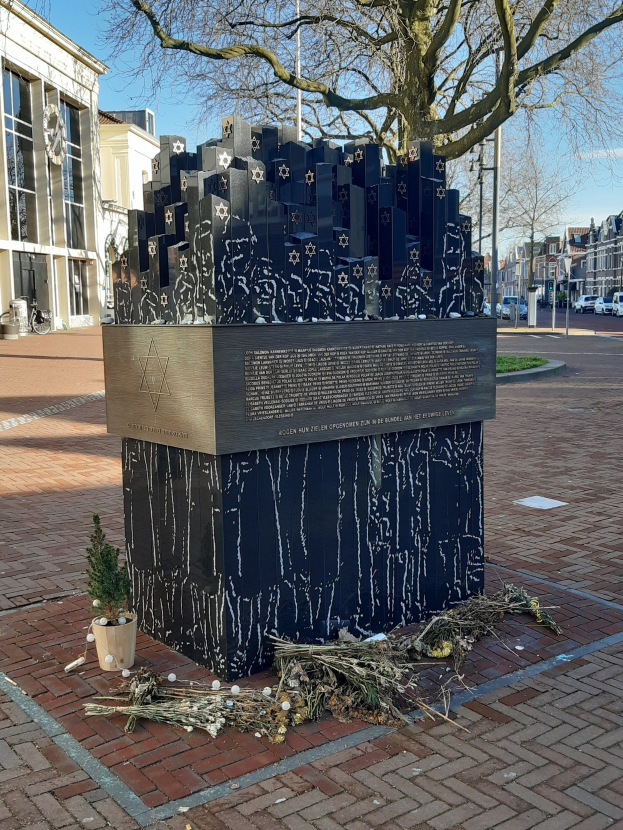 Denkmal für Holocaust-Opfer in einem Stadtplatz mit einem Baum davor, einem Topf mit einer Pflanze auf dem Boden, verstreuten trockenen Blättern und Gebäuden, Fahrzeugen, Masten und Bäumen im Hintergrund.