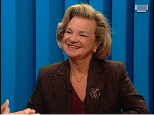 Eine strahlende Frau in einer braunen Jacke mit einem Dutt, die Hände verschränkt, vor einem blauen Hintergrund.