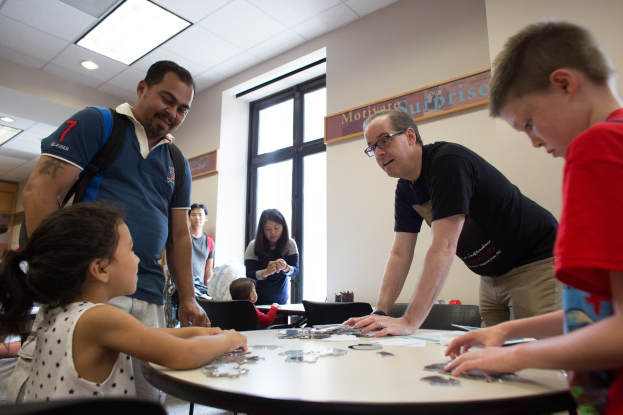 Gruppe von Menschen an einem Tisch mit Puzzles, einem Fenster in der Wand, einer Tafel mit Schrift und Deckenbeleuchtung im Hintergrund.