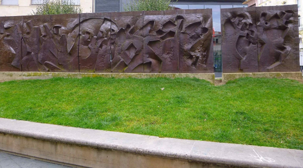Eine große Metallskulptur, die die Berliner Mauer darstellt, steht auf einer grasbewachsenen Fläche, umgeben von Bäumen und Gebäuden mit Fenstern im Hintergrund.