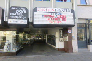 Das Lincoln Theater in Berlin, Deutschland, ein Gebäude mit Glasfenstern und -türen und einer Tafel mit Text darauf, mit verschiedenen Gegenständen im Inneren, die einen pulsierenden Stadtbild Eindruck vermitteln.