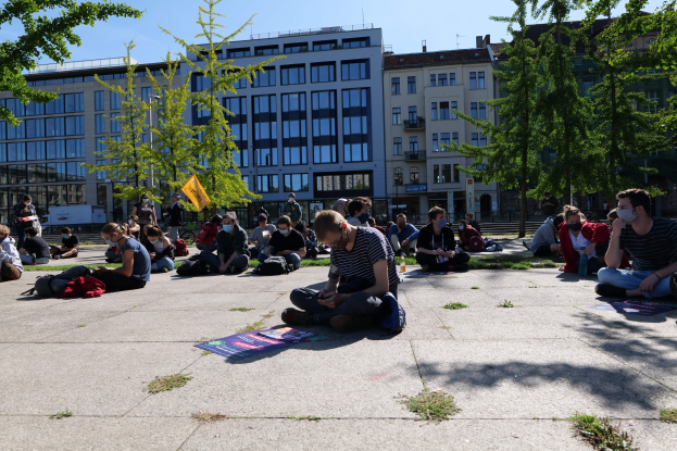 Eine Gruppe von Menschen sitzt vor einem Gebäude auf dem Boden während einer Demonstration in Berlin, einige tragen Masken, mit verstreuten Taschen und Gegenständen, umgeben von Bäumen unter einem klaren blauen Himmel.