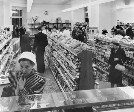 Schwarzes und weißes Foto von Menschen beim Einkaufen in einem Supermarkt mit Lebensmittelregalen, einige halten Taschen, eine Uhr an der Wand und Deckenbeleuchtung.