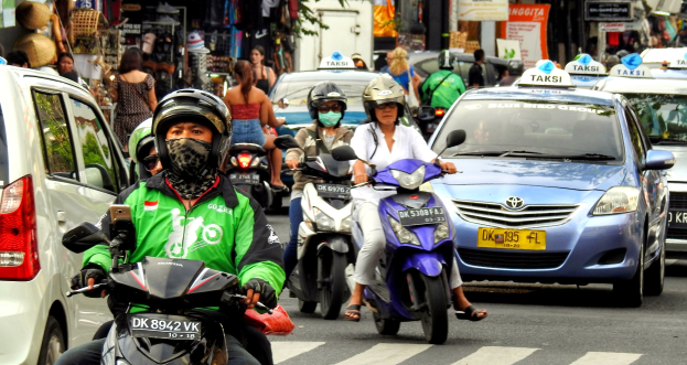 Eine Gruppe von Menschen auf Motorrädern fährt auf einer belebten Straße mit Autos, Gebäuden, Bäumen und Geschäften im Hintergrund.
