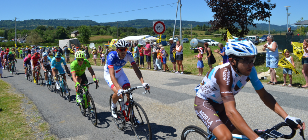Gruppe von Radfahrern auf einer Straße mit Zuschauern an den Seiten, einige tragen Helme und Schutzbrillen oder halten Fahnen, vor einem Hintergrund aus Bäumen, Schildern, Fahrzeugen, Zelten, Gebäuden, Hügeln und einem klaren blauen Himmel.
