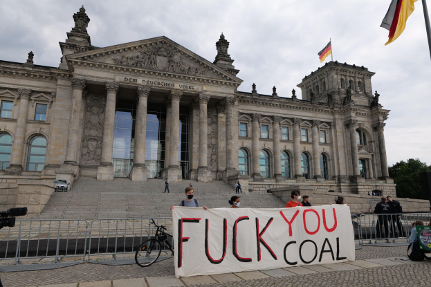 Eine Gruppe von Menschen mit einem "Fuck You Coal"-Schild vor dem Reichstag in Berlin, mit Bäumen, Fahnenmast und bewölktem Himmel im Hintergrund.